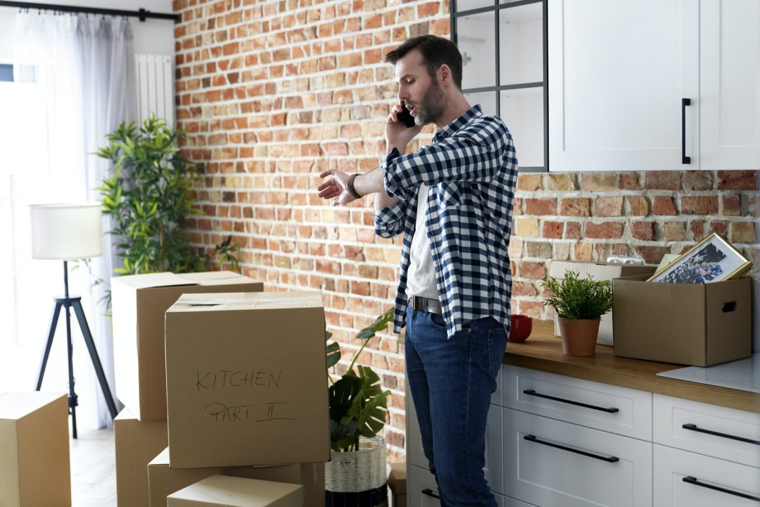 A man in a checkered shirt speaks on the phone while standing among stacked moving boxes in a modern kitchen with a brick wall.