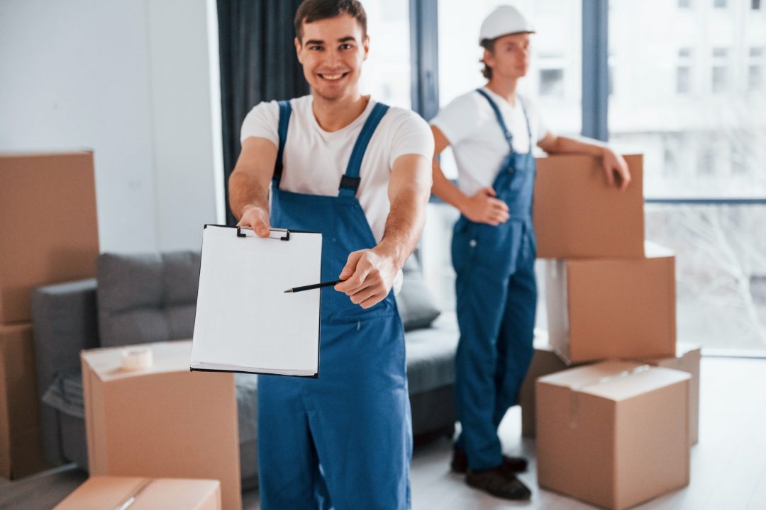 Two movers in blue overalls are inside a room filled with cardboard boxes. One mover is holding a clipboard and smiling, while the other stands in the background, also smiling.