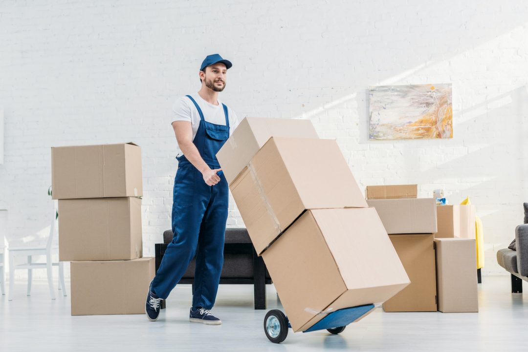 A mover in overalls pushes a dolly stacked with cardboard boxes in a bright, spacious room. Several boxes are arranged nearby, indicating an ongoing move.