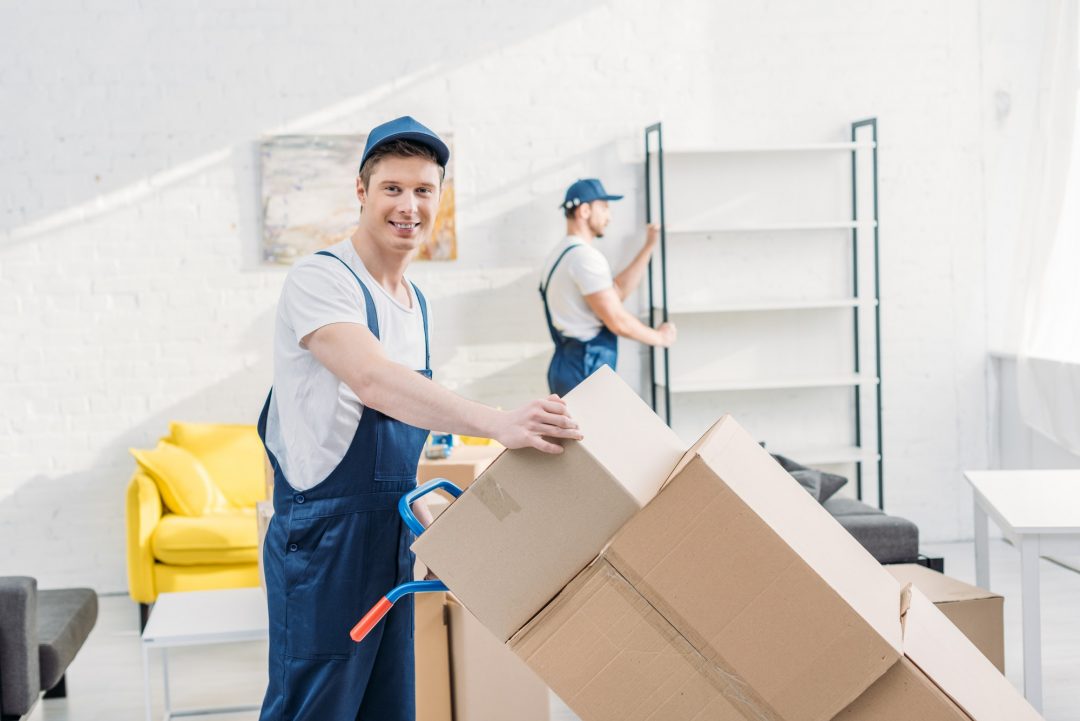 Two movers are packing boxes in a bright, modern room. One mover is smiling while lifting a box, and another is organizing shelves in the background.