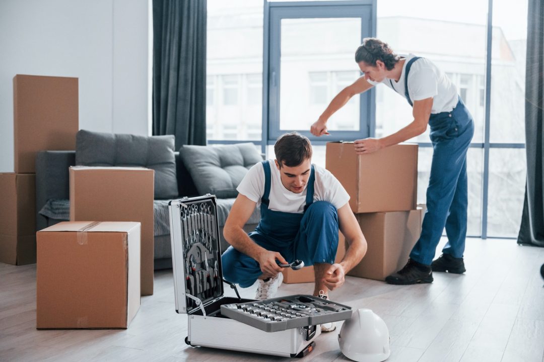 Two movers are in a bright room with large windows. One is kneeling next to a disassembled appliance, while the other is standing and packing boxes.